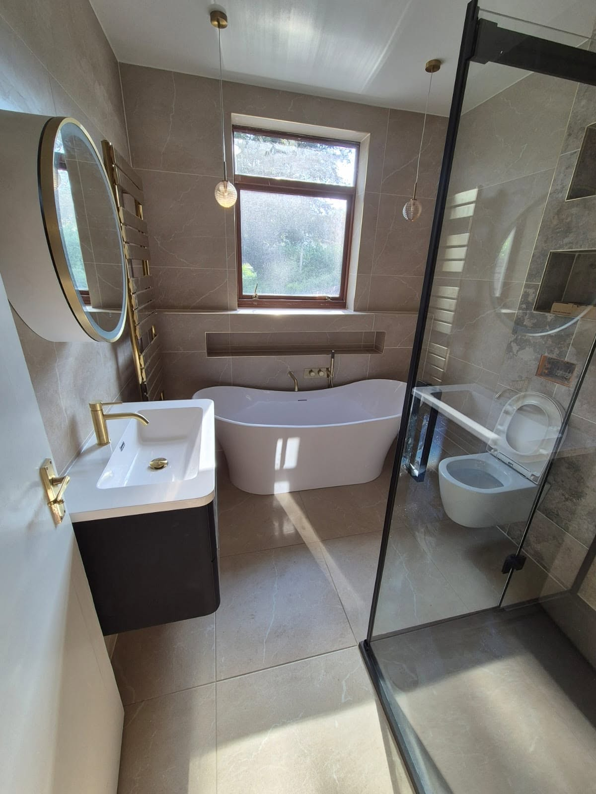 New bathroom with warm marble tiling, freestanding bath, gold brassware and round LED mirror